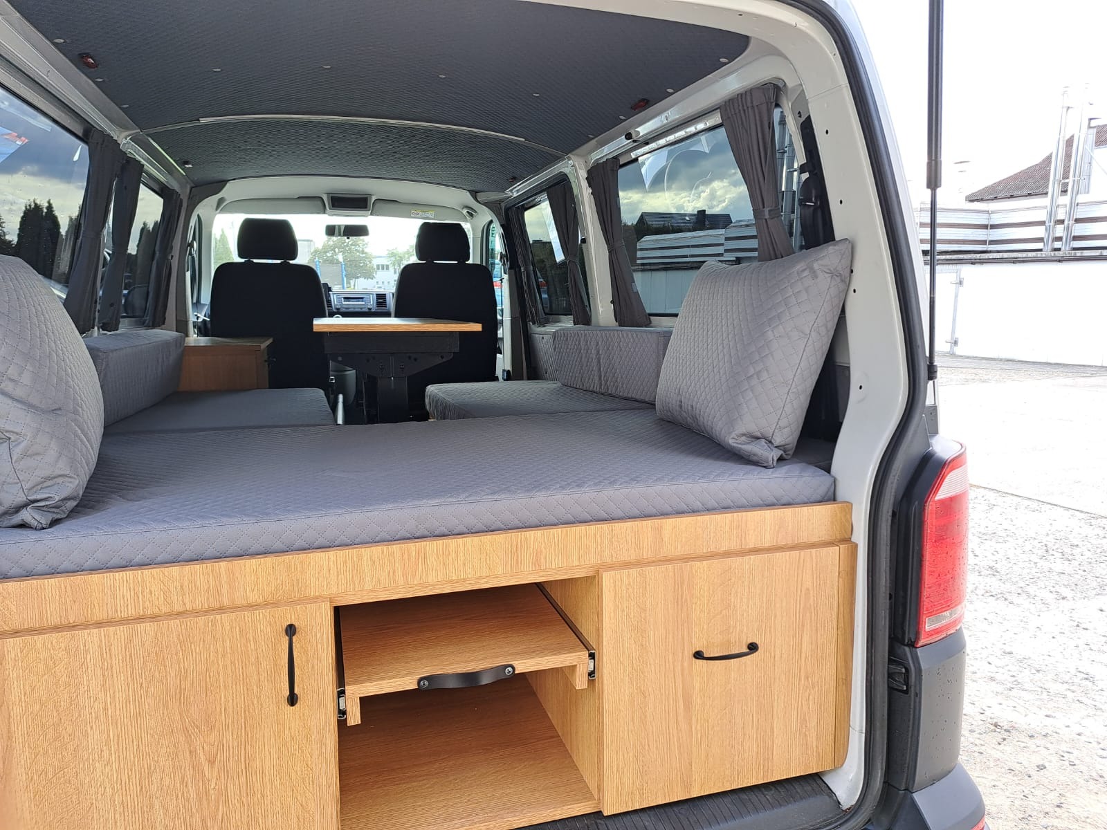 Zabudowa kampera z drewnianymi szafkami, szarą tapicerką i rozkładanym stolikiem. Widok z tyłu pojazdu, wnętrze jasne, z zasłonkami w oknach.