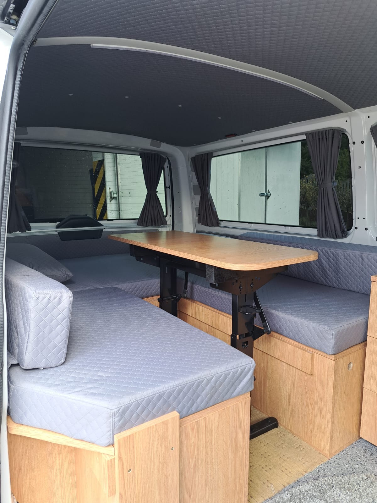 Wnętrze kampera z drewnianą zabudową: szare tapicerowane siedziska, regulowany stół. Widoczne zasłony w oknach i pikowana podsufitka. Funkcjonalne rozwiązanie.