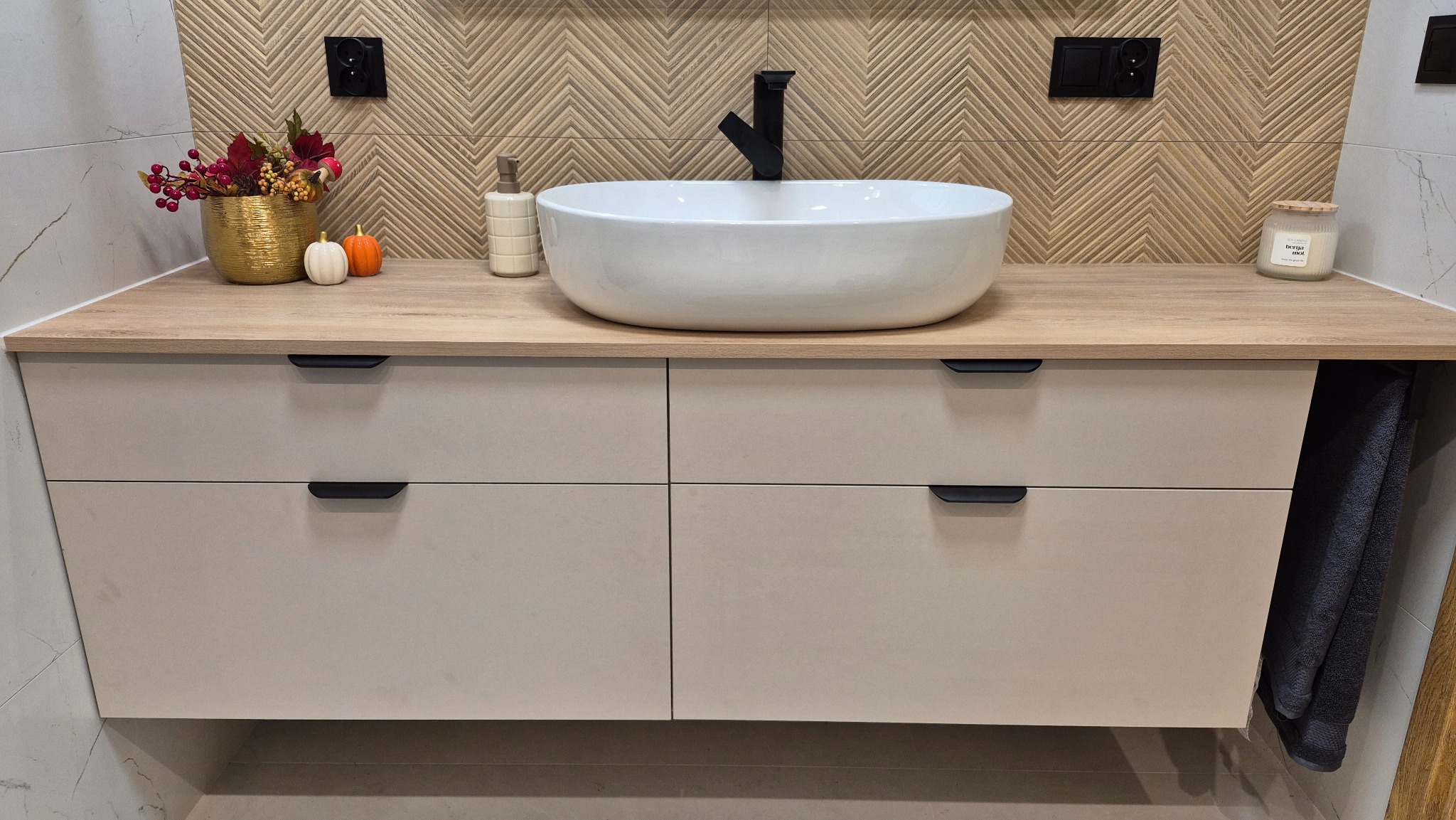Nowoczesna szafka łazienkowa z blatem imitującym drewno, białą umywalką i czarną armaturą. Na blacie dekoracje: świeca, dozownik i złoty wazon z jesiennymi kwiatami.