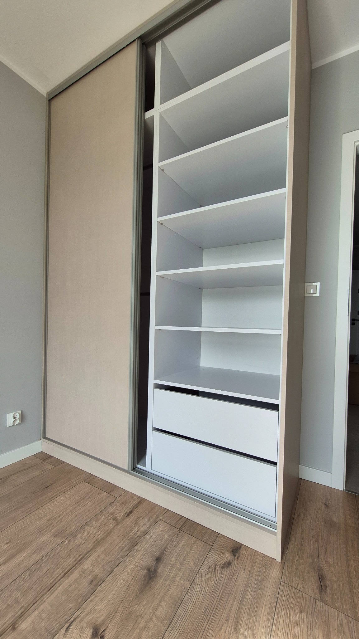 Jasna szafa wnękowa z przesuwnymi drzwiami i białymi półkami, widoczna na tle drewnianej podłogi i szarej ściany. Minimalistyczny design, nowoczesne wykończenie.
