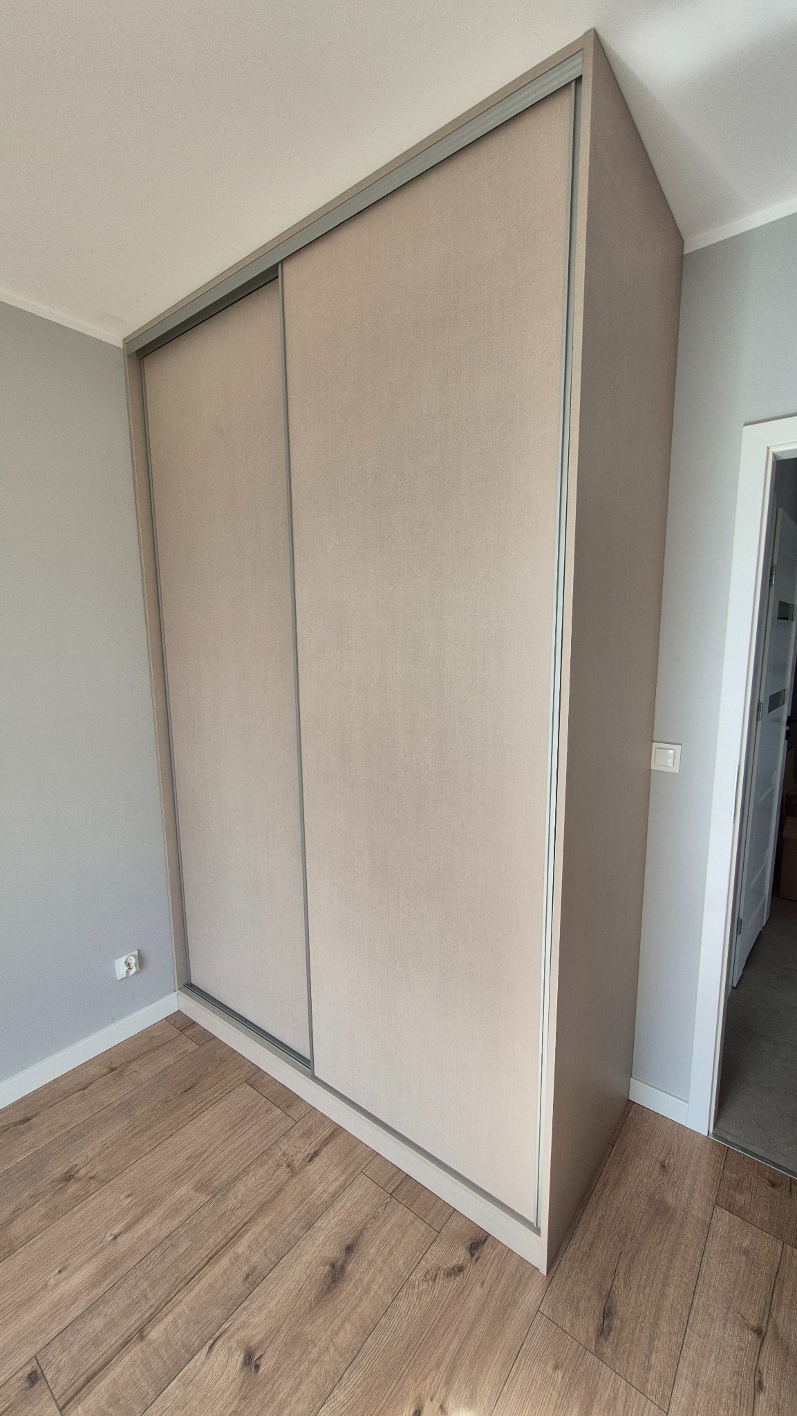 Beżowa szafa wnękowa z przesuwnymi drzwiami, wykończona srebrnymi detalami, na tle drewnianej podłogi i szarej ściany. Widok z perspektywy narożnika pokoju.