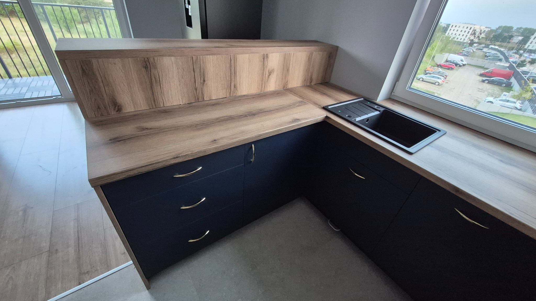 Ciemnoniebieska zabudowa kuchenna z drewnianym blatem i złotymi uchwytami, zlew wpuszczony w blat przy oknie z widokiem na parking.