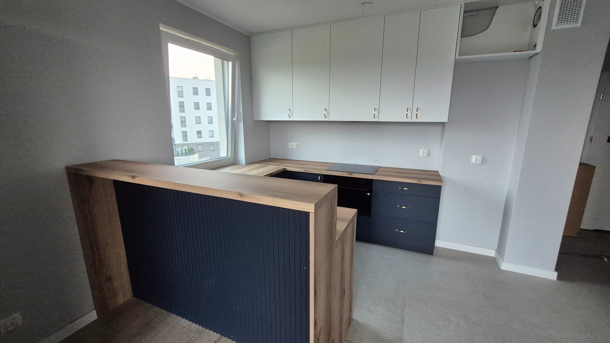 Nowoczesna kuchnia z barkiem: białe szafki wiszące, ciemne dolne szafki z drewnianym blatem, okno z widokiem na budynek. Minimalistyczny design.