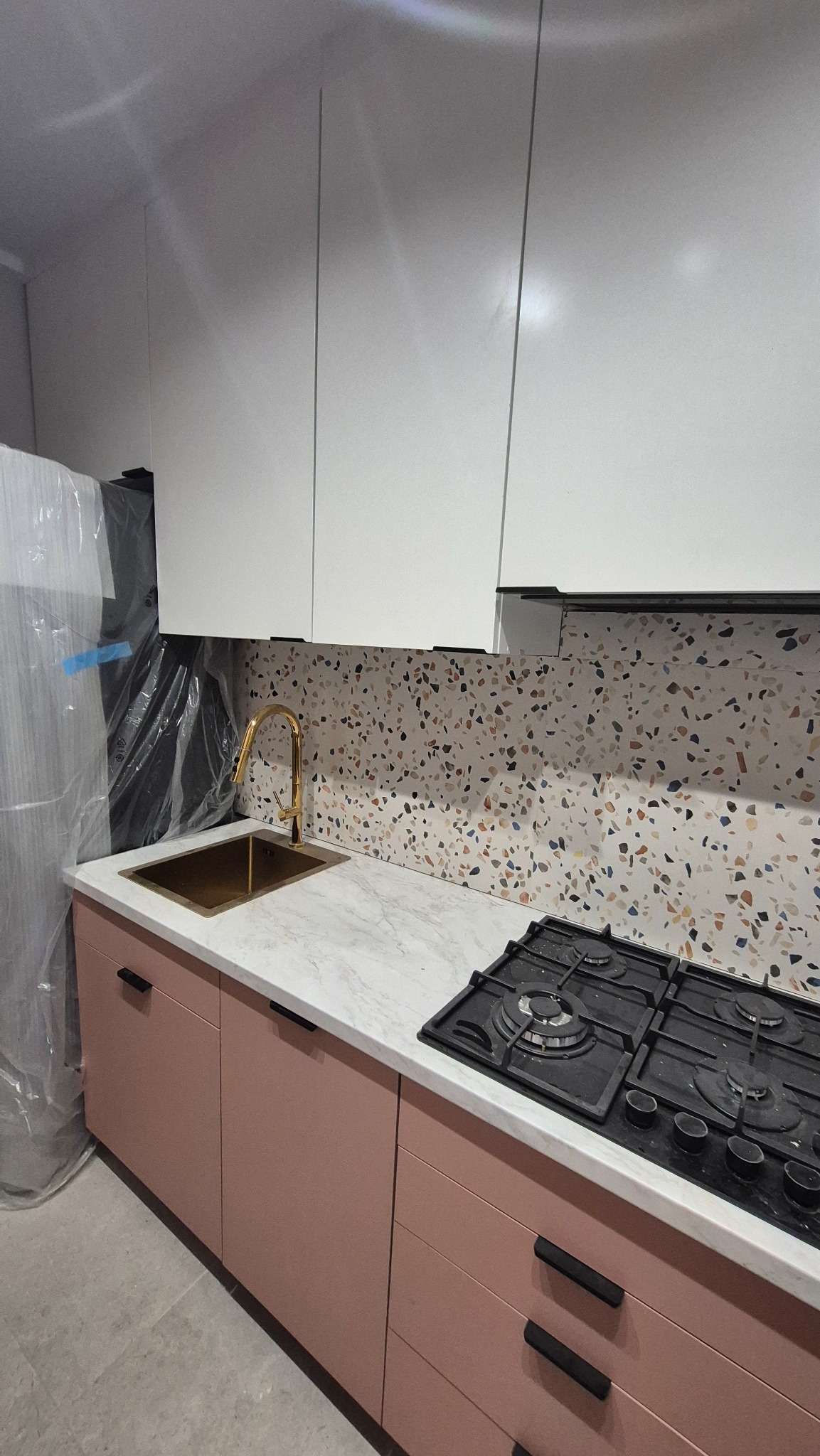 Nowoczesna kuchnia z różowymi szafkami, marmurowym blatem i złotą armaturą. Na blacie czarna płyta gazowa. Ściana wyłożona mozaiką w stylu lastryko.