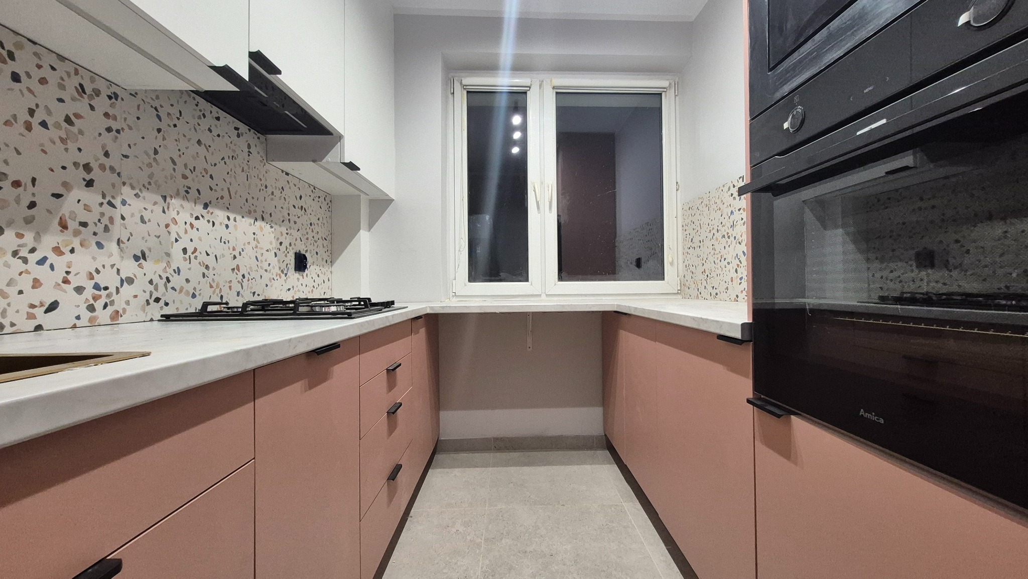 Różowa zabudowa kuchenna z białym blatem i mozaiką na ścianie, widok na okno i piekarnik Amica. Nowoczesny design z czarnymi uchwytami.