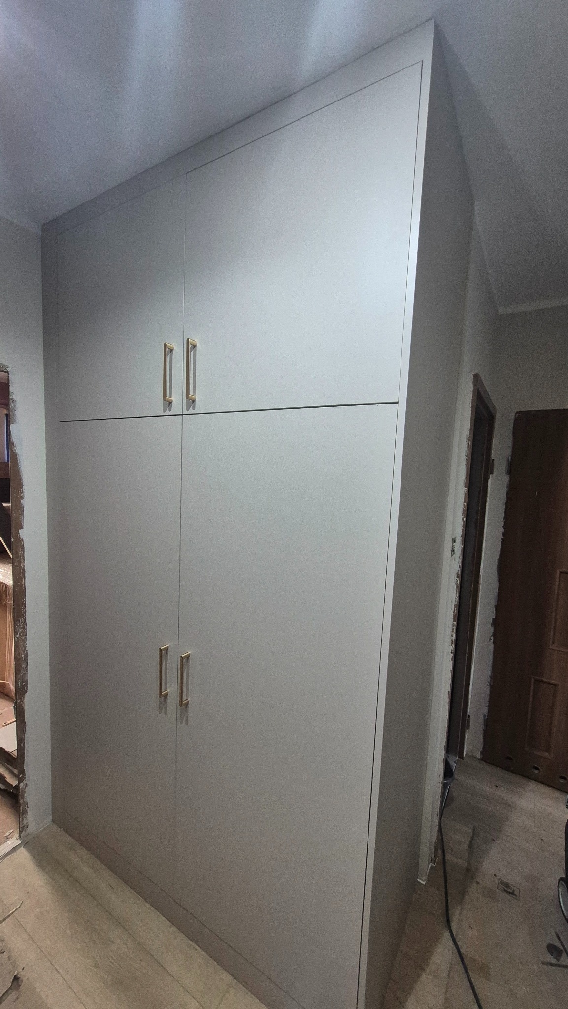 Szara szafa wnękowa z złotymi uchwytami, dopasowana do skosu sufitu, widoczna w trakcie montażu, z fragmentem niezabezpieczonej ściany w tle.