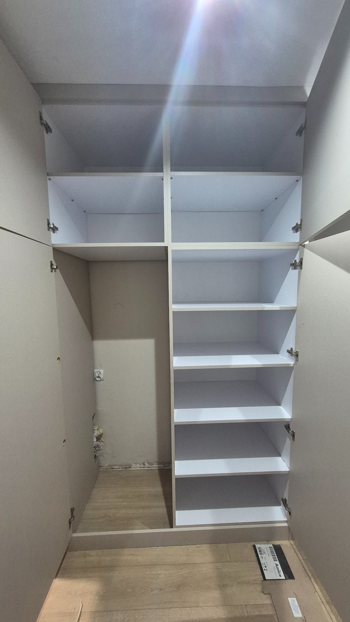 Wnętrze szafy wnękowej z białymi półkami, widoczne zawiasy i fragment ściany z gniazdkiem elektrycznym. Ujęcie z perspektywy wnętrza szafy.