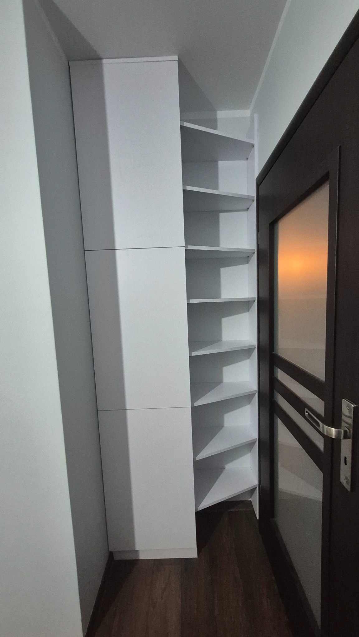 Biała szafa narożna z półkami, wkomponowana we wnękę obok drzwi. Minimalistyczny design, gładkie fronty, widoczne półki w kształcie trójkątów. Funkcjonalne rozwiązanie do przechowywania.