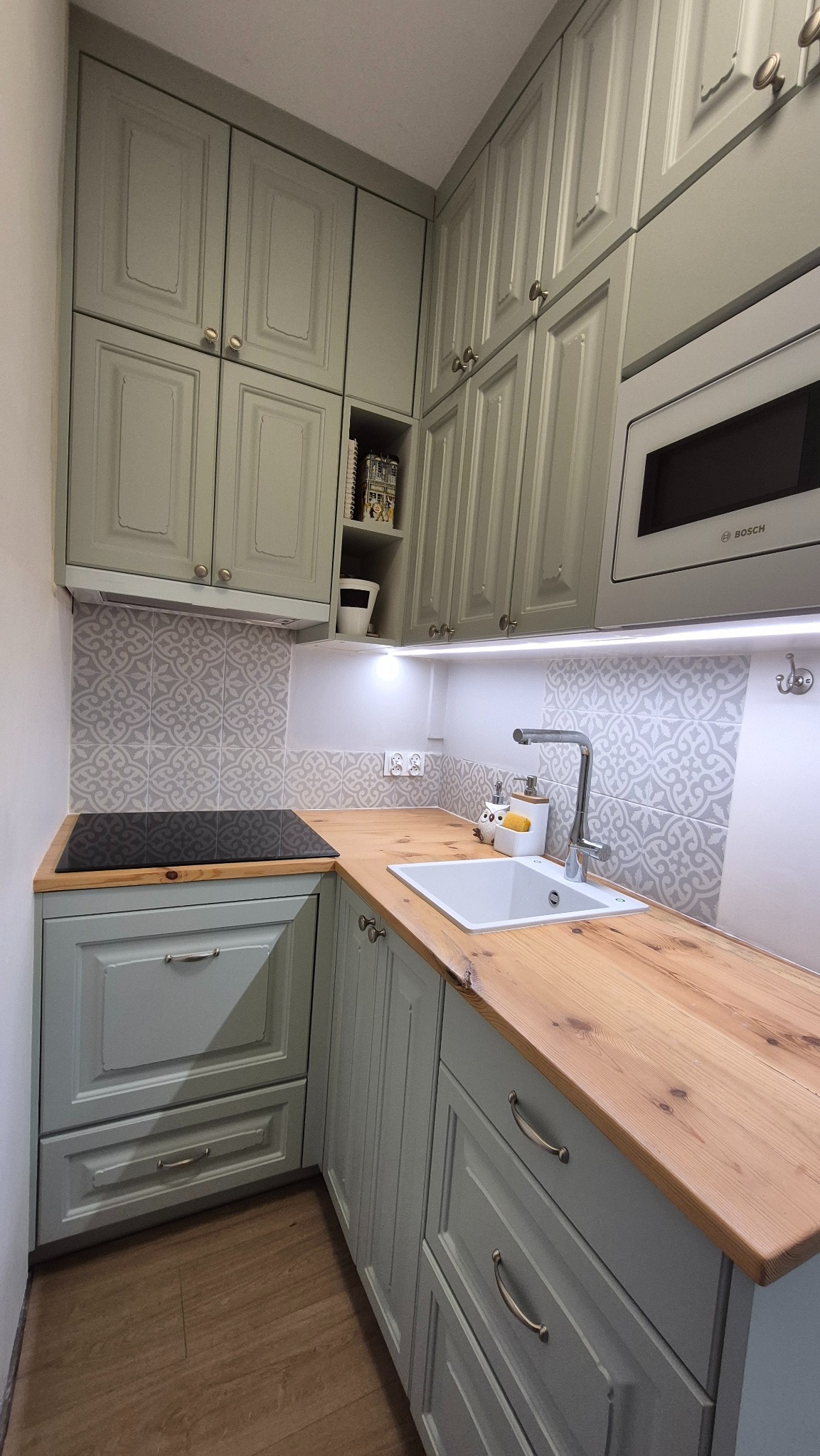 Jasnoszara kuchnia na wymiar z drewnianym blatem i wzorzystą mozaiką nad blatem. Widoczna płyta indukcyjna i zlew. Oświetlenie LED podszafkowe.