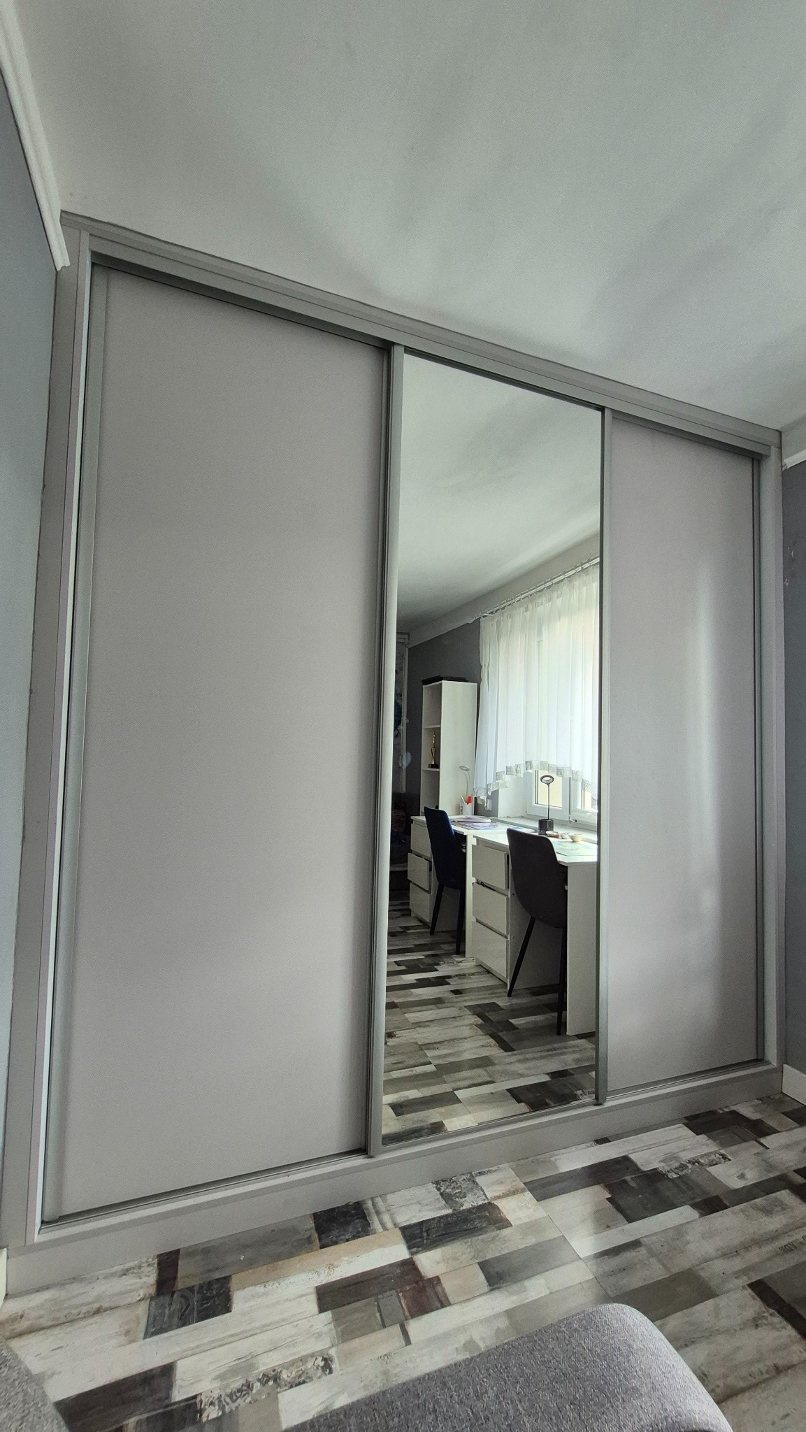 Jasnoszara szafa wnękowa z przesuwnymi drzwiami i lustrem, odbijająca pokój z biurkiem i krzesłami. Podłoga z paneli w odcieniach szarości i brązu.