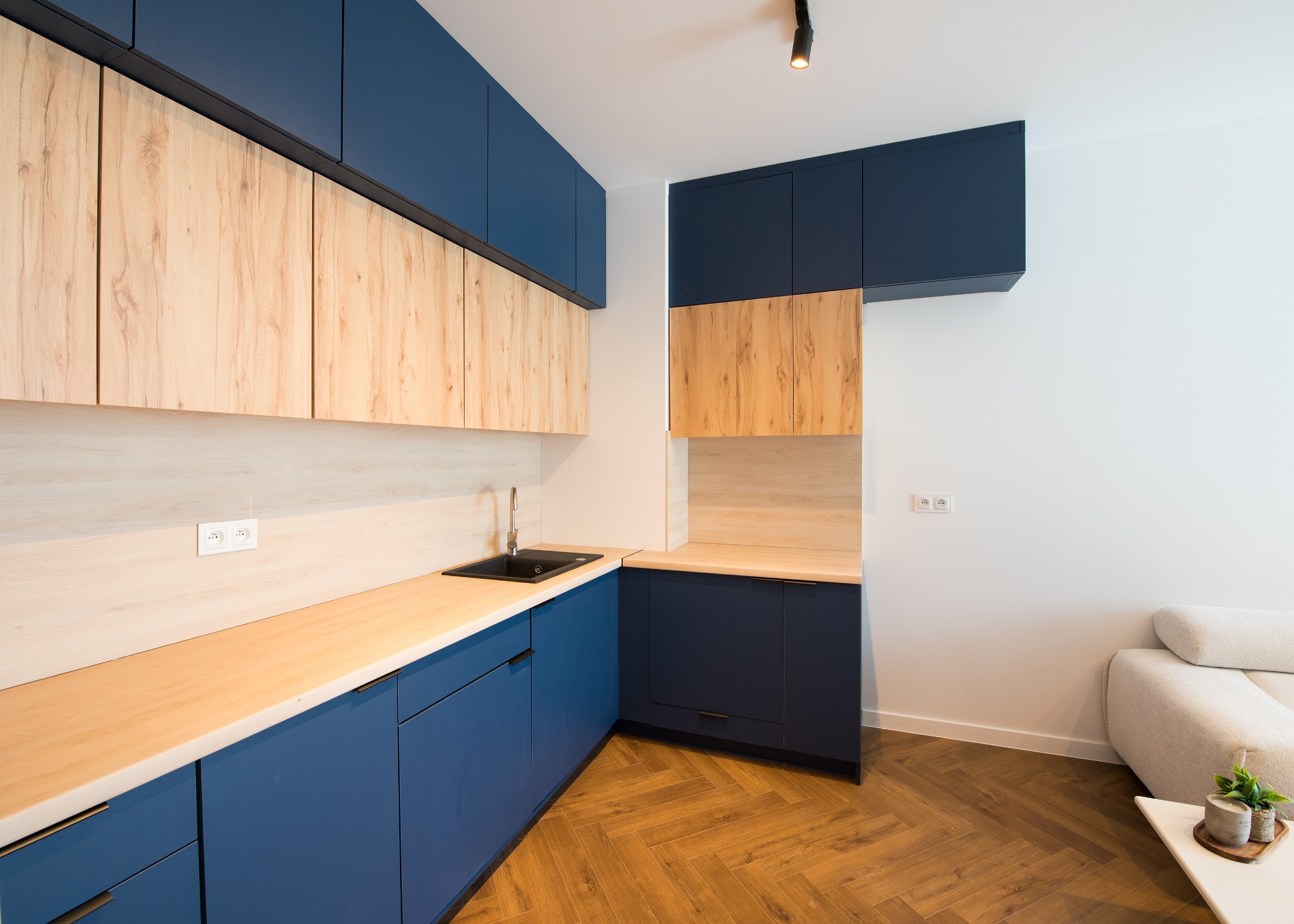 Niebiesko-drewniana kuchnia z czarnym zlewem i parkietem w jodełkę. Górne szafki z drewnianymi frontami, dolne i wiszące szafki w kolorze granatowym. Nowoczesny design.