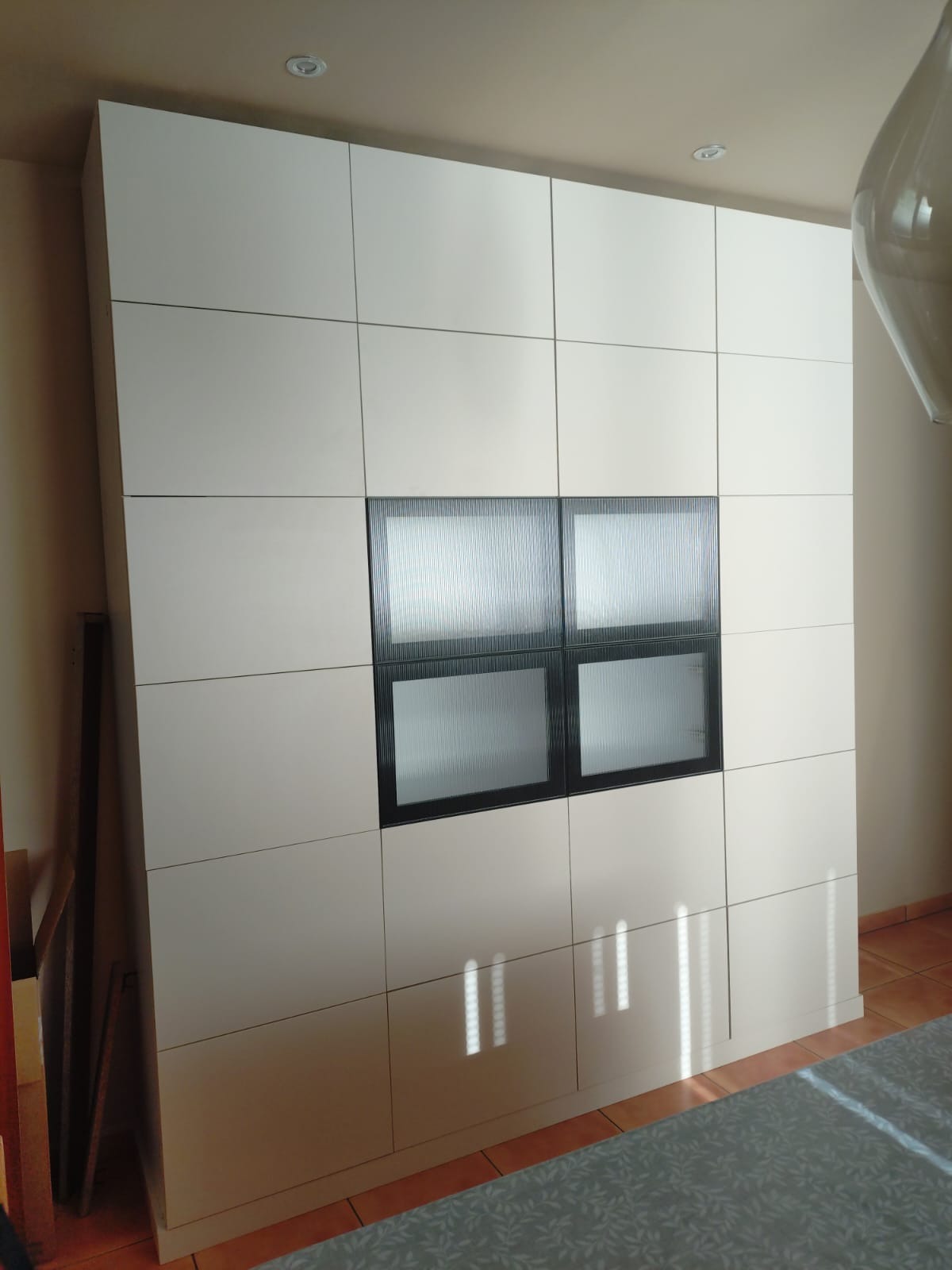 Biała szafa wnękowa z kwadratowymi panelami i czterema przeszklonymi szybami z ryflowanego szkła, tworzącymi okno w centralnej części.