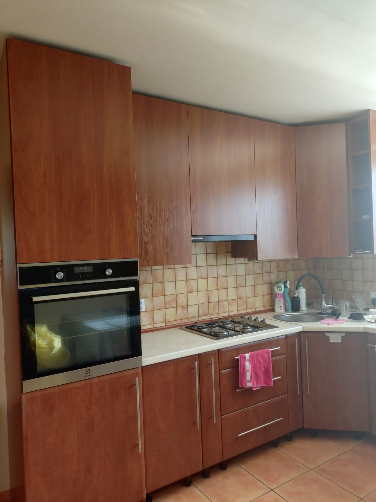 Brązowa zabudowa kuchenna z piekarnikiem Electrolux i płytą gazową. Beżowe płytki na ścianie. Różowy ręcznik na szafce. Ujęcie z bliska, widok na fragment kuchni.