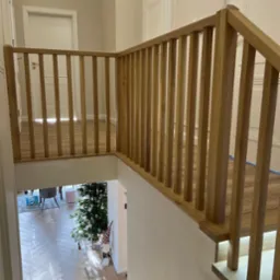 Jasna, drewniana balustrada z pionowymi szczebelkami, widok z góry na salon z parkietem w jodełkę i choinką, minimalistyczne wnętrze.