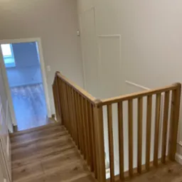 Klatka schodowa z drewnianą balustradą i panelami podłogowymi imitującymi drewno, widok z góry na dół, jasne wnętrze z białymi ścianami.