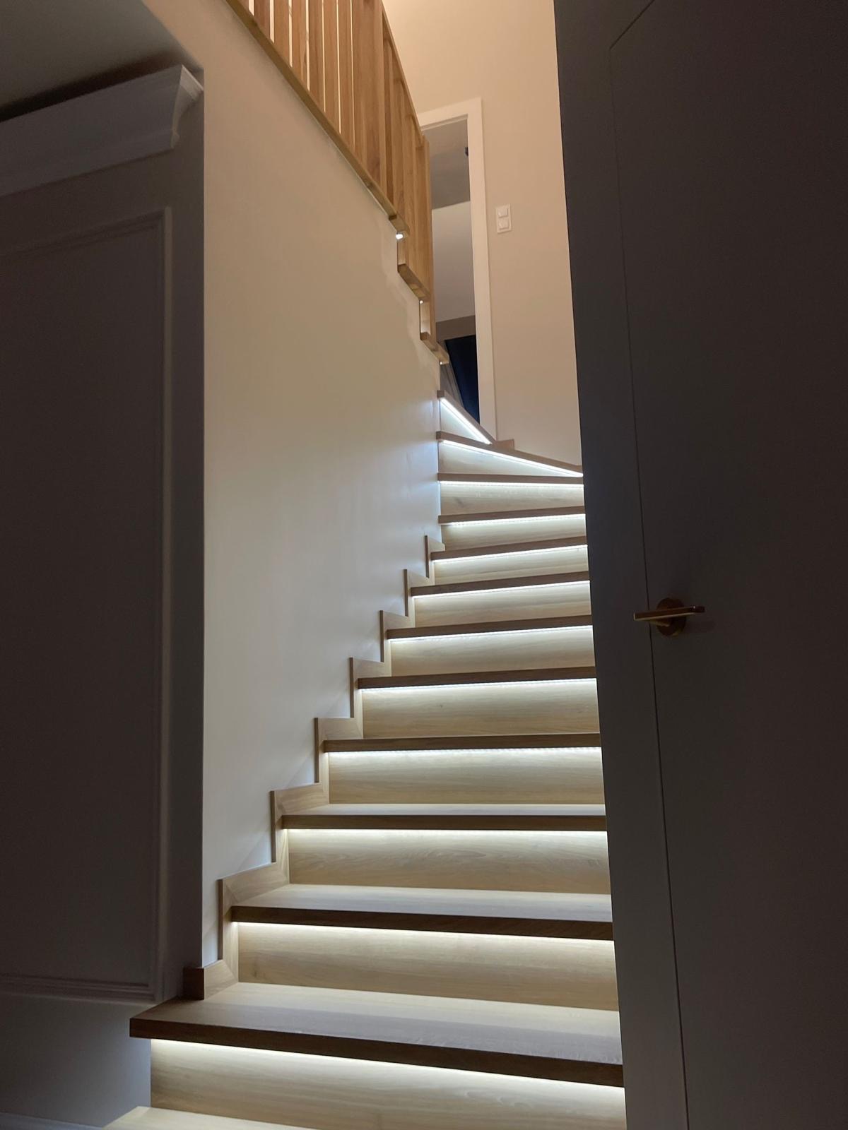 Klatka schodowa z podświetlanymi stopniami, drewniana balustrada, widok z dołu. Stopnie oświetlone ledowymi paskami, jasne drewno, minimalistyczny design.