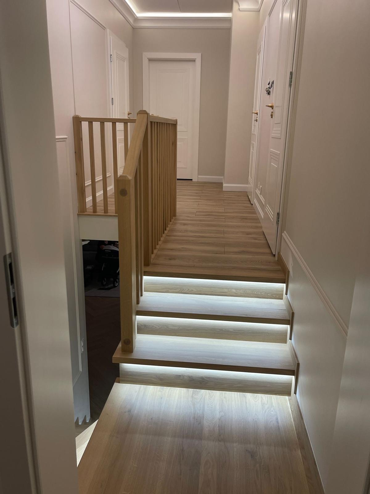 Jasne drewniane schody z podświetleniem LED zamontowanym pod każdym stopniem, prowadzące na wyższy poziom domu. Balustrada z jasnego drewna po lewej stronie schodów. W tle widoczne białe drzwi...