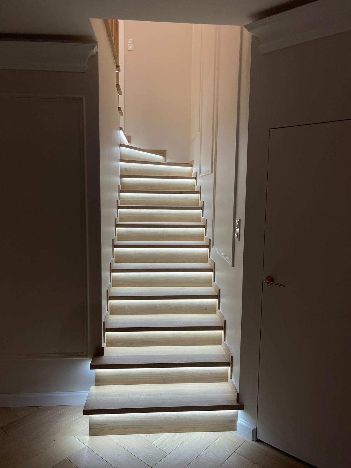 Nowoczesne schody z podświetleniem LED, widok z dołu, jasne drewno, minimalistyczny design, drzwi po prawej stronie.