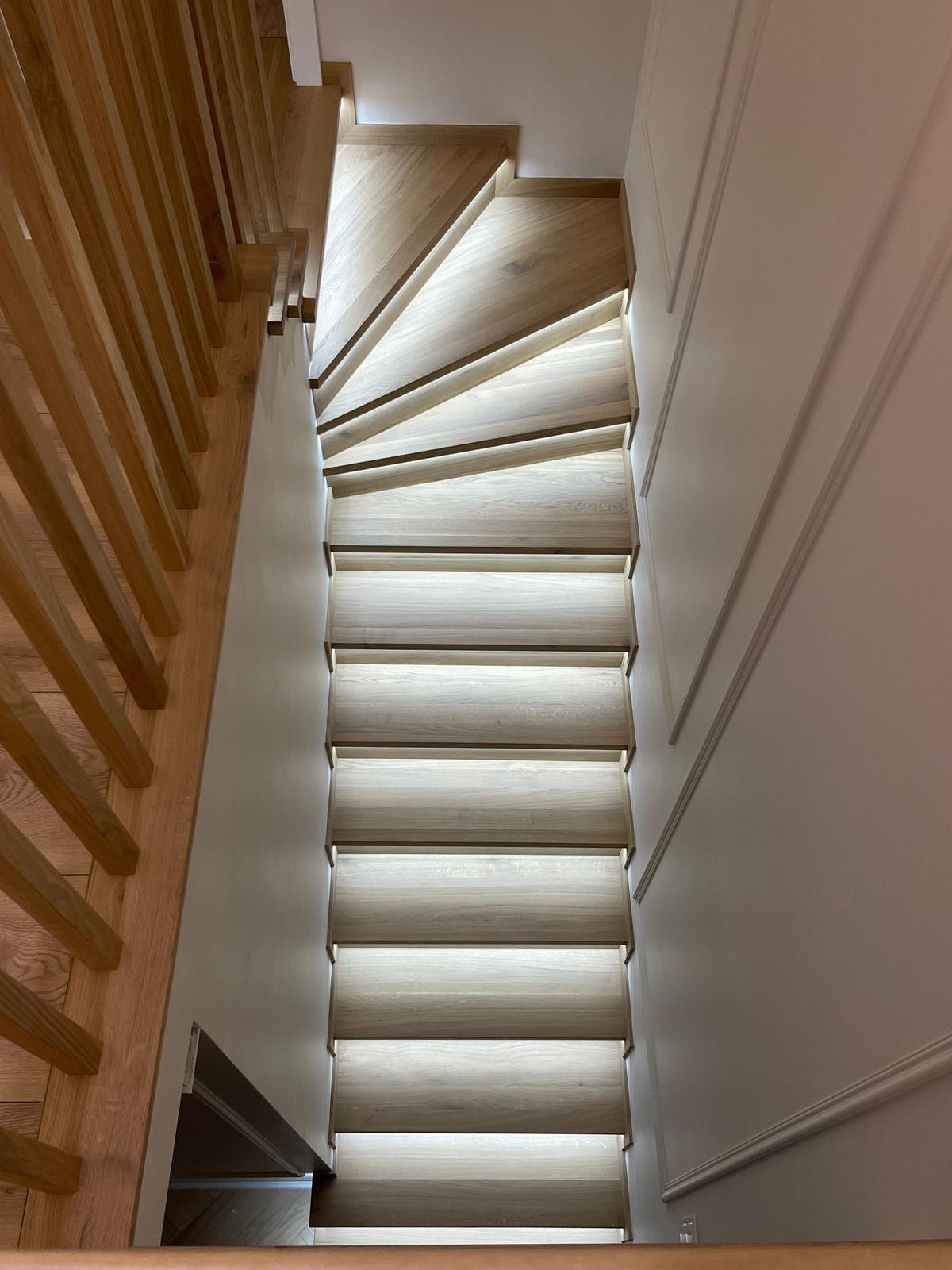 Wewnętrzne schody z jasnego drewna z podświetleniem LED, widok z góry, z drewnianą balustradą po lewej i ozdobną sztukaterią na ścianie po prawej.