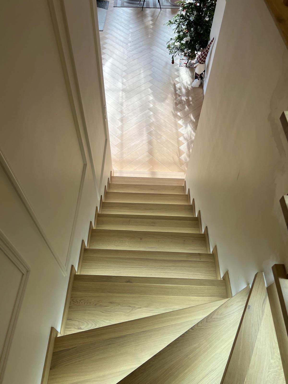 Widok z góry na jasne, dębowe stopnie schodów z podświetleniem LED, prowadzące do pomieszczenia z parkietem w jodełkę i choinką w tle.