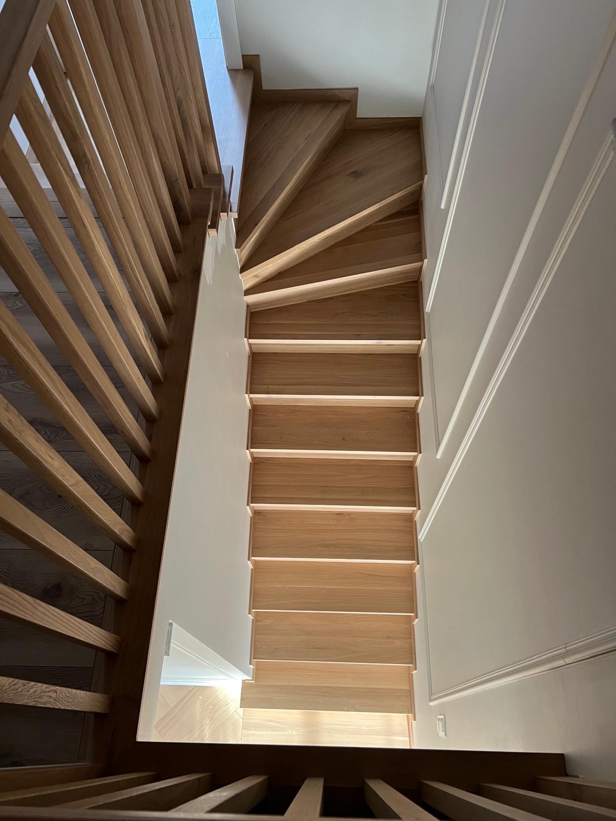 Wysokie ujęcie krętych schodów drewnianych z balustradą z jasnego drewna, kontrastujące z białą ścianą ozdobioną sztukaterią.