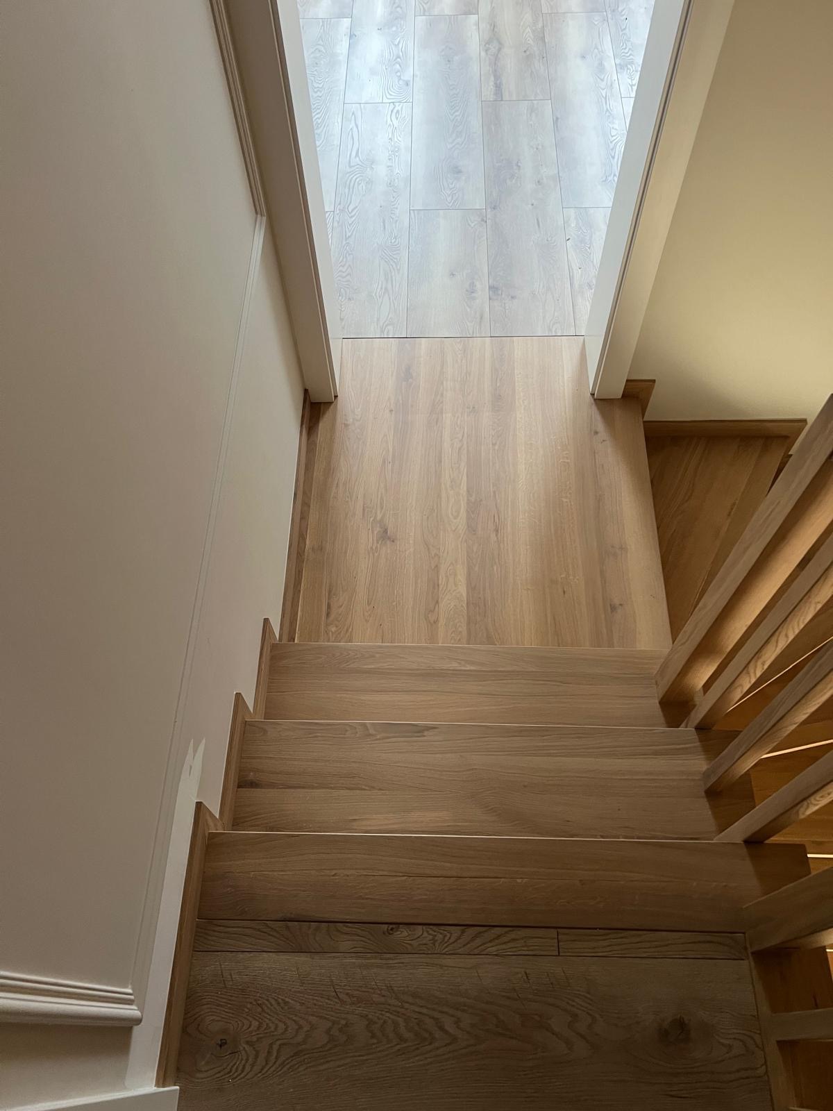 Widok z góry na dębowe schody z drewnianą balustradą, prowadzące na jasną podłogę, z białymi ścianami po bokach.