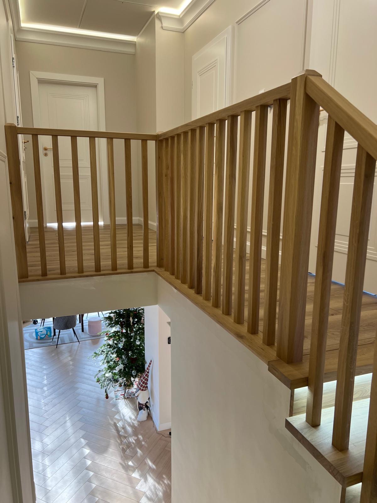 Jasna, drewniana balustrada z pionowymi szczebelkami, widok z góry na salon z parkietem w jodełkę i choinką, minimalistyczne wnętrze.