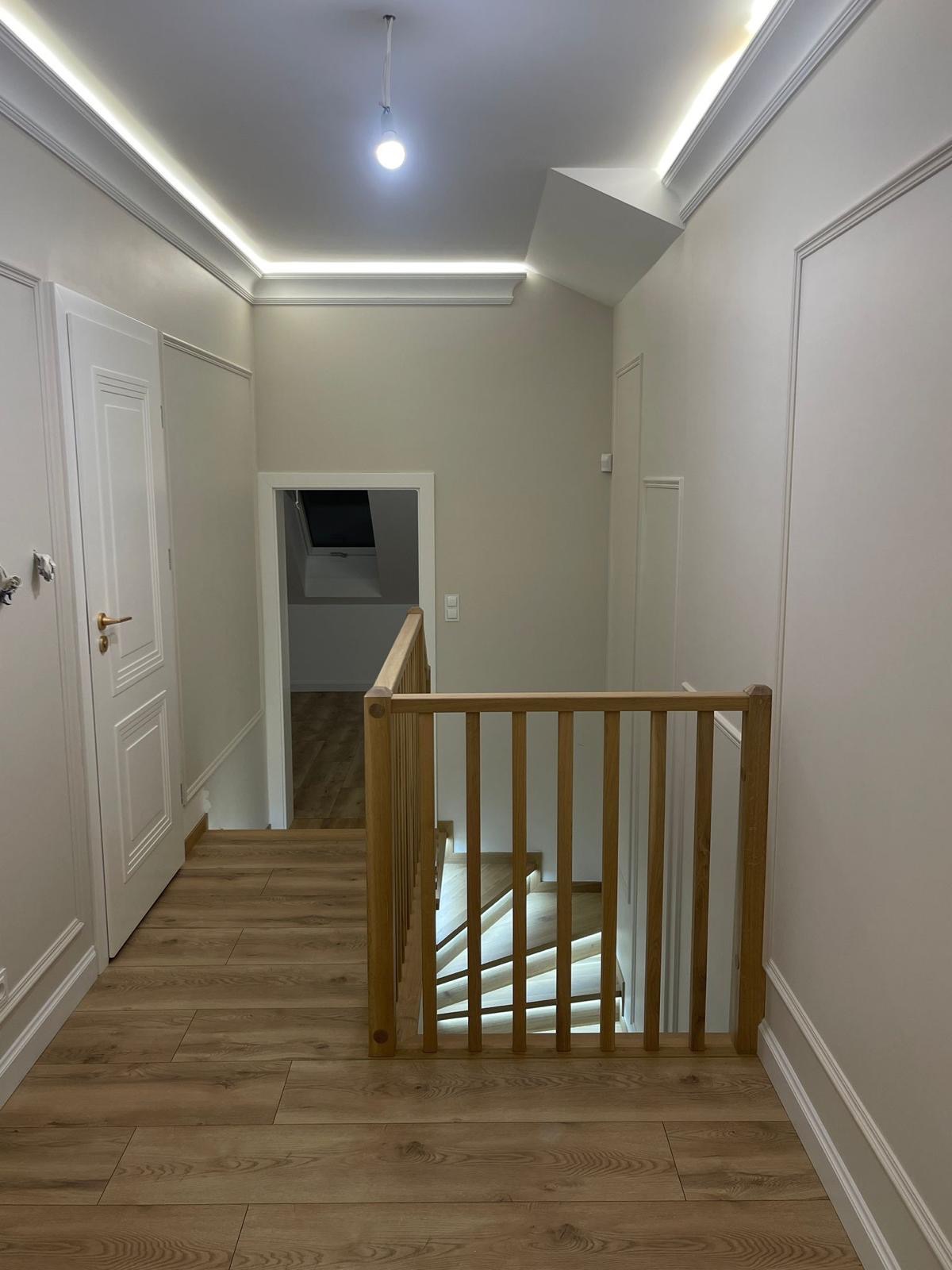 Widok z góry na jasne, drewniane schody z balustradą, oświetlone od spodu, w nowoczesnym wnętrzu z kremowymi ścianami i drewnianą podłogą.