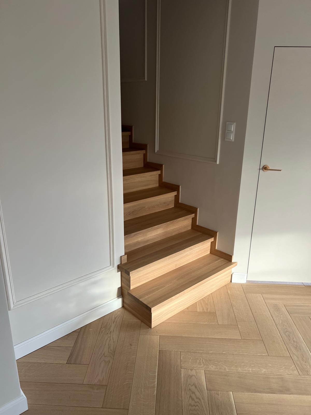 Jasne, drewniane schody zintegrowane z podłogą w jodełkę, minimalistyczne wykończenie ścian z dekoracyjnymi listwami.