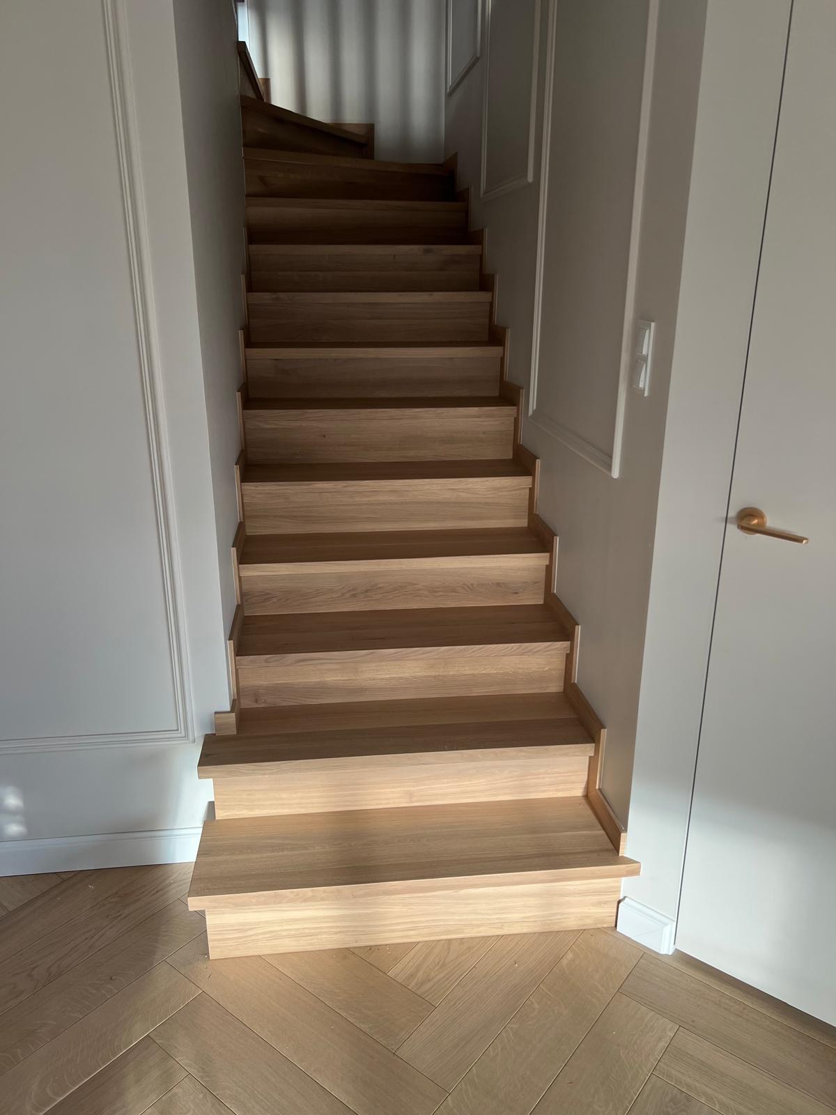 Jasne, dębowe stopnie schodowe bez balustrady, z widocznym wzorem jodełki na podłodze i białymi panelami ściennymi.