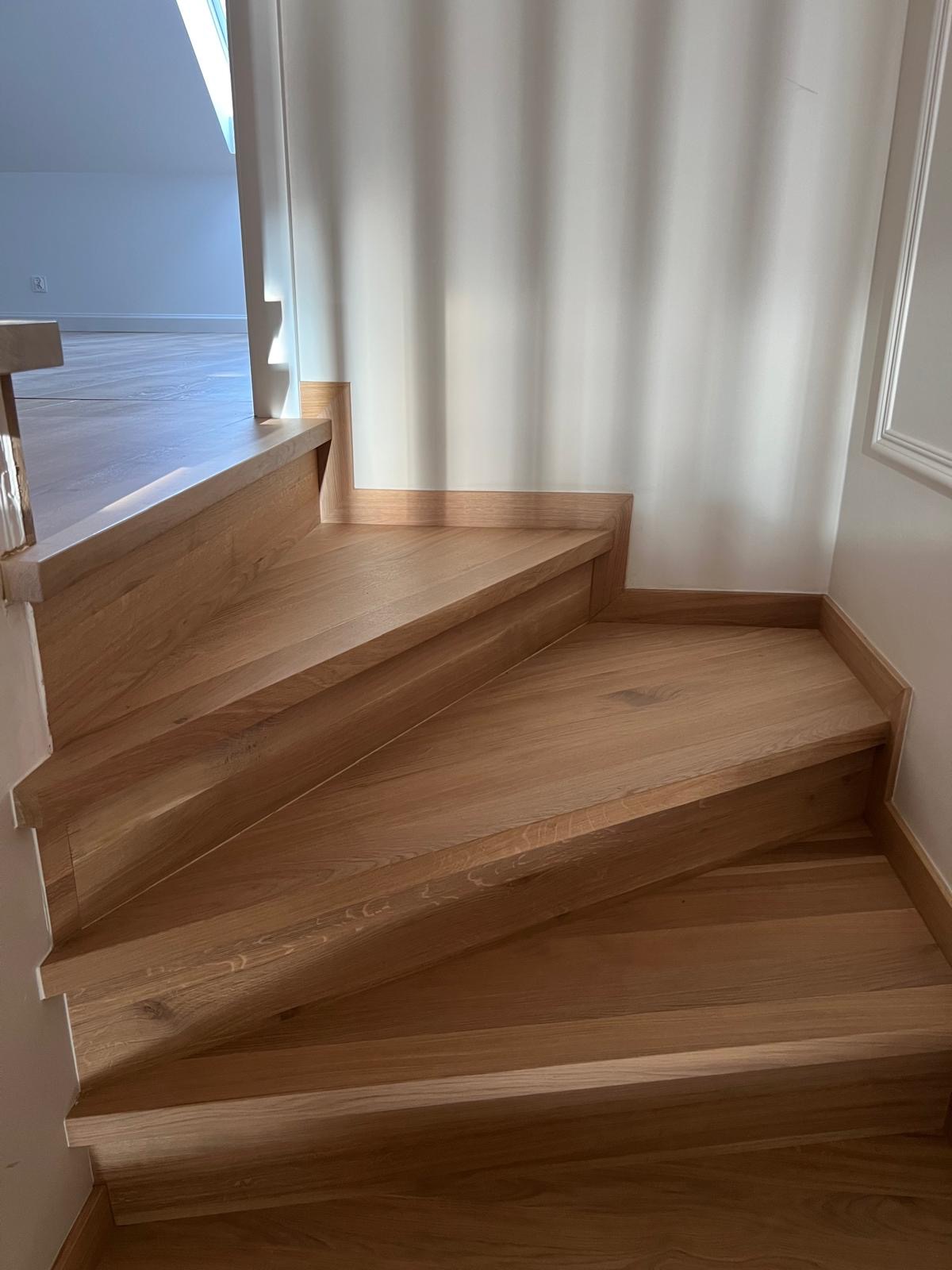 Jasne, dębowe stopnie schodowe bez balustrady, z widoczną strukturą drewna i delikatnym oświetleniem padającym z góry.