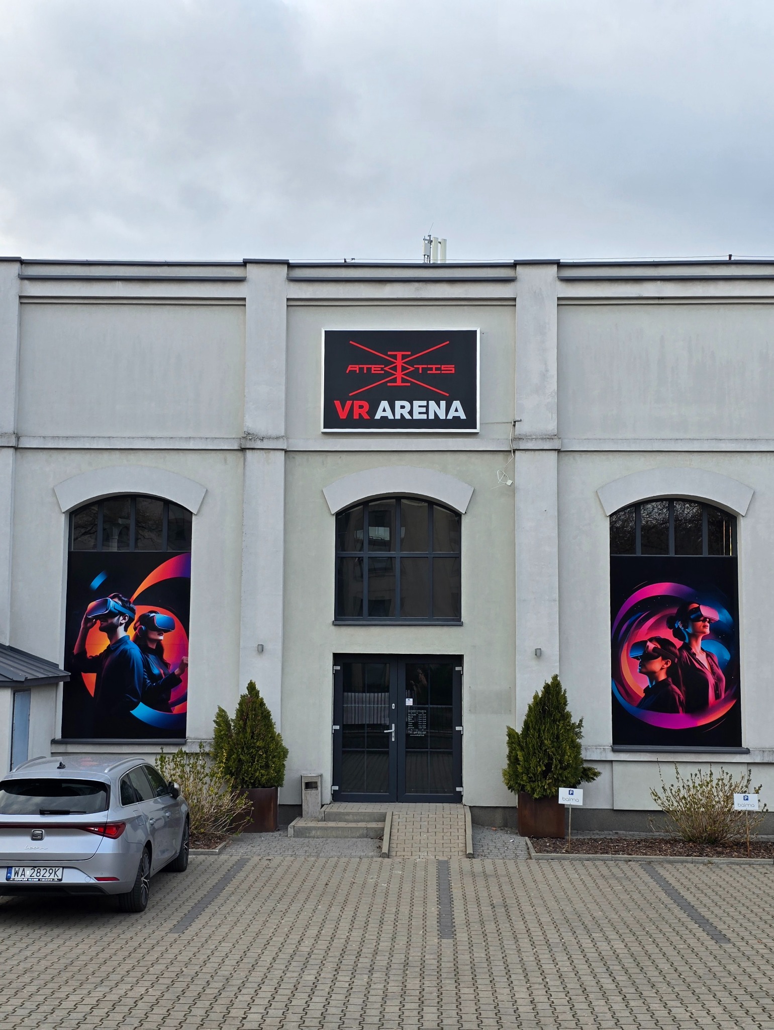 Fasada budynku VR Arena z logo, plakatami przedstawiającymi osoby w goglach VR, szary samochód zaparkowany z boku.