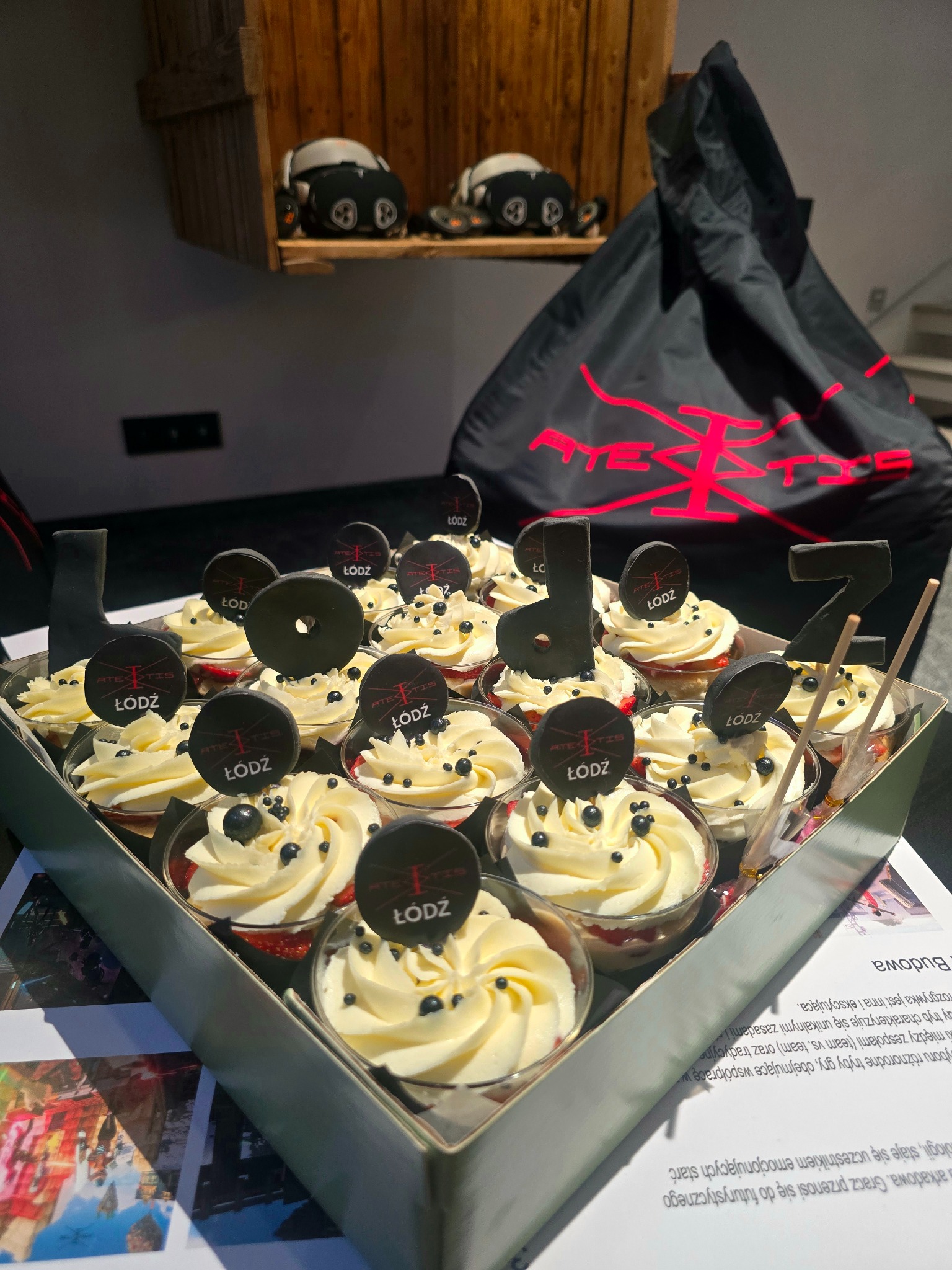 Deserki w przezroczystych pucharkach z kremem, czarnymi kuleczkami i logiem 'Łódź' oraz czarne cyfry '1' i '7' w metalowej tacy, w tle torba z czerwonym logo firmy.