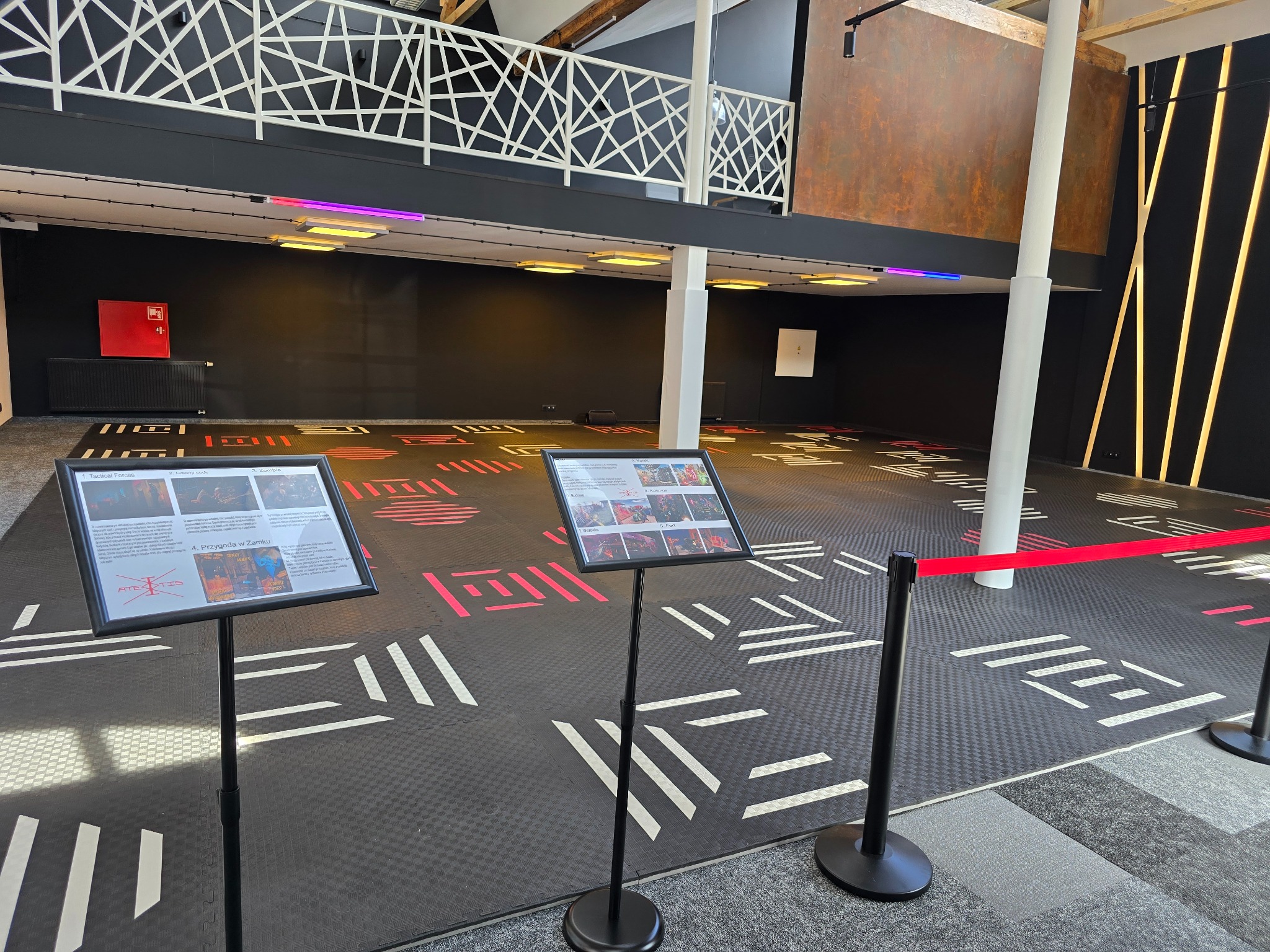 Sala gier z podłogą w geometryczne wzory, dwa stojaki z informacjami o atrakcjach, czerwona taśma oddzielająca przestrzeń, oświetlenie LED w kolorach fioletu i żółci.