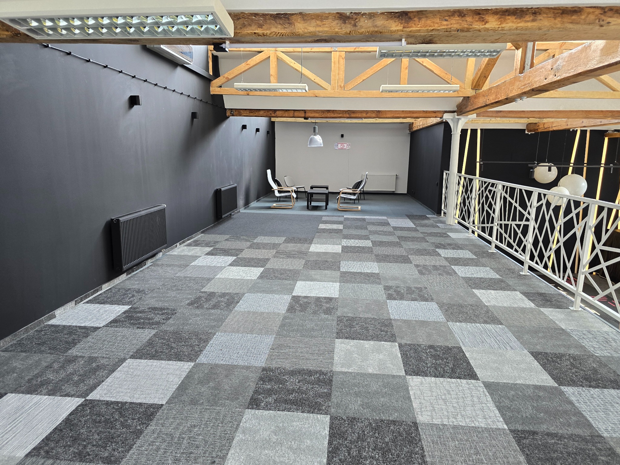Przestronne, nowoczesne biuro z szarą wykładziną w kratę, czarnymi ścianami i drewnianymi belkami stropowymi, oświetlone lampami sufitowymi. W głębi widoczne minimalistyczne meble wypoczynkowe.