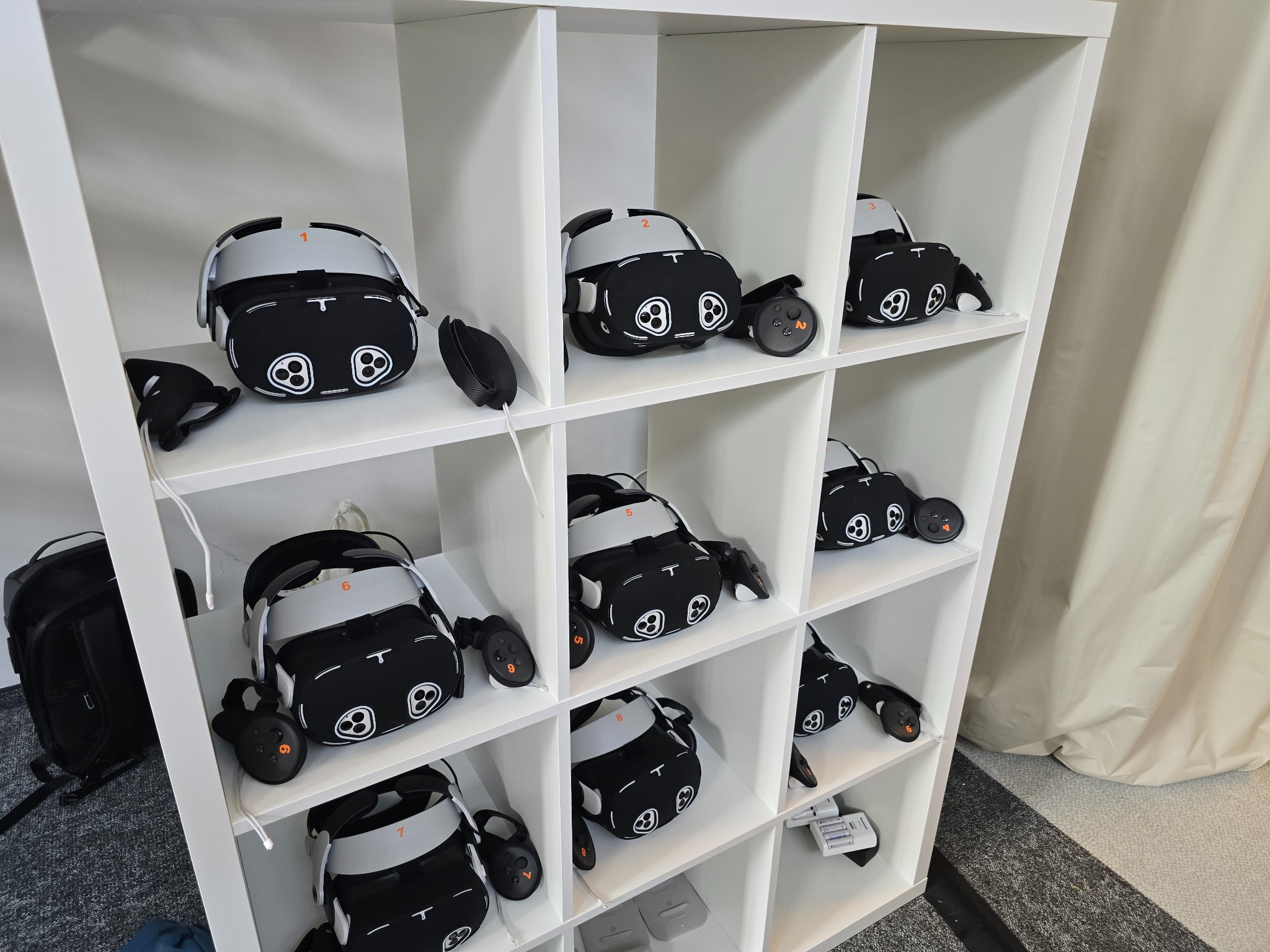 Regał z ponumerowanymi zestawami VR (gogle i kontrolery) przygotowanymi do użytku.