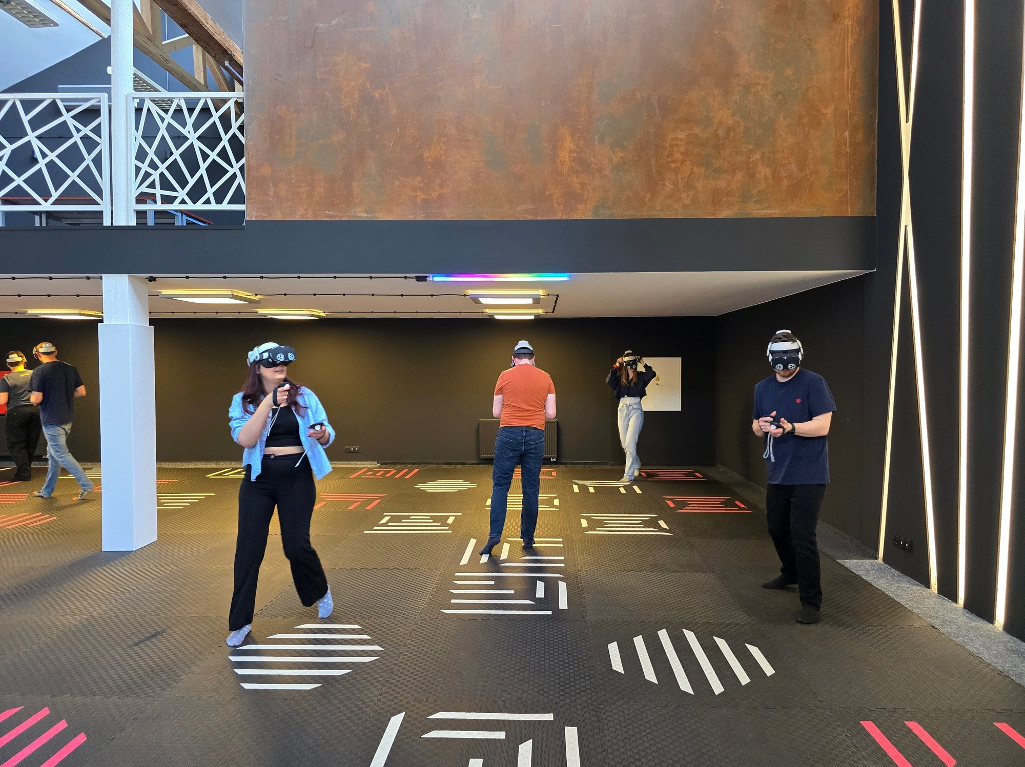 Grupa osób z goglami VR w przestronnym pokoju z czarnymi ścianami i podłogą z geometrycznymi wzorami. Oświetlenie LED. Atrakcja na imprezę firmową.