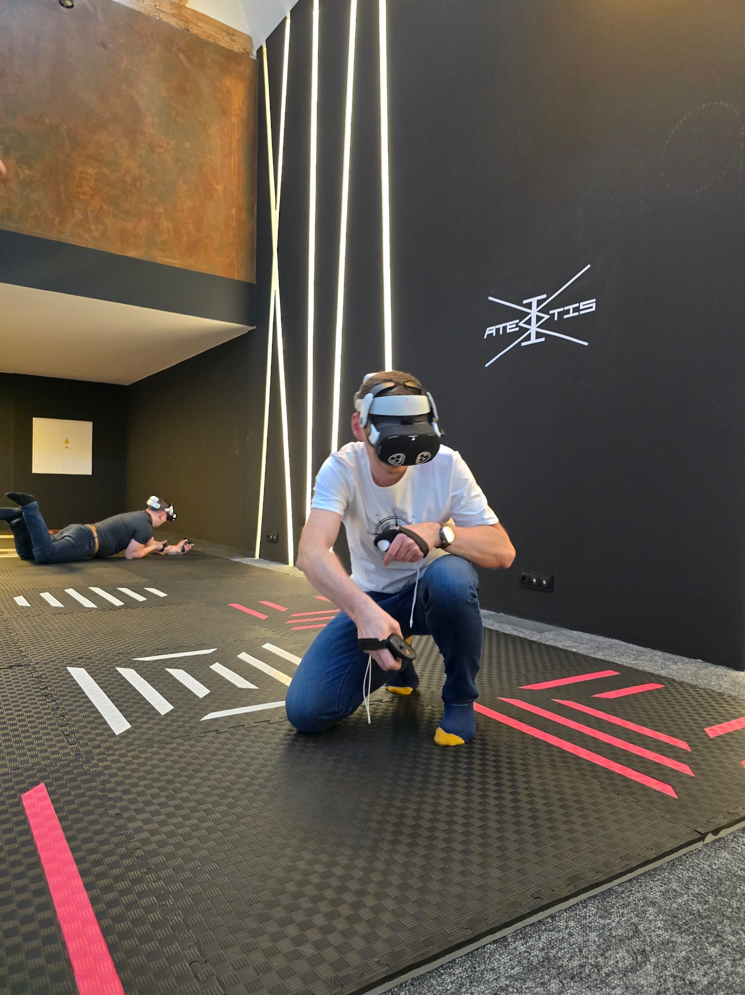 Gracze VR w akcji: mężczyzna w goglach VR kuca na interaktywnej podłodze, drugi gracz leży w tle. Dynamiczna rozrywka w przestrzeni eventowej.