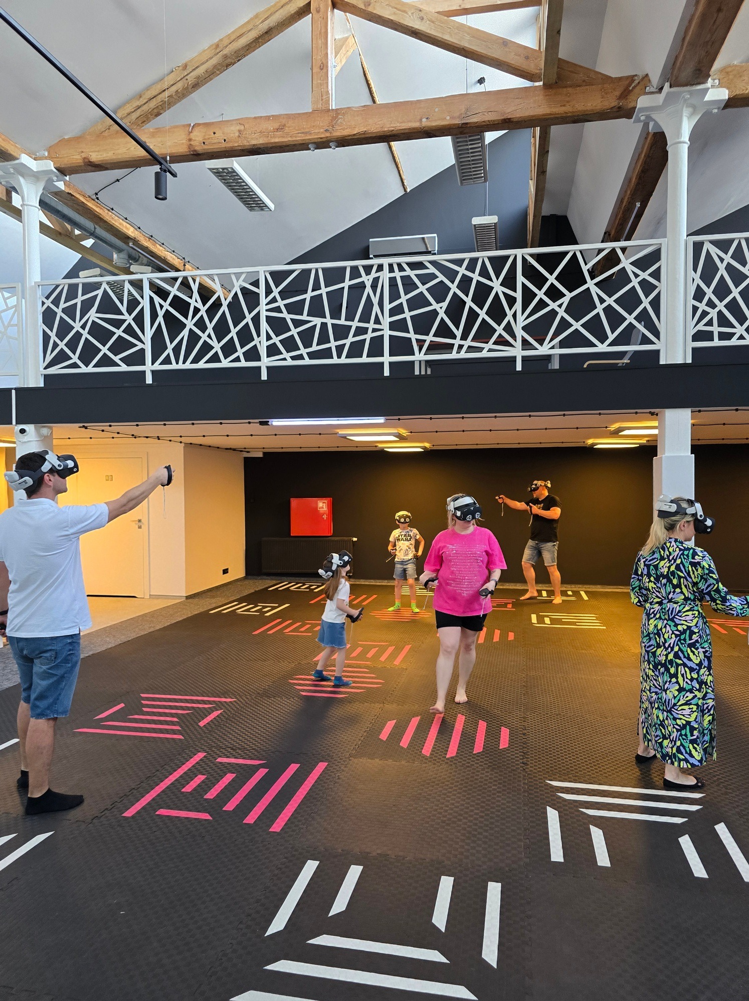 Grupa osób w goglach VR w przestronnym, industrialnym wnętrzu z podłogą w geometryczne wzory, uczestnicząca w interaktywnej rozrywce. Widoczne drewniane belki stropowe.
