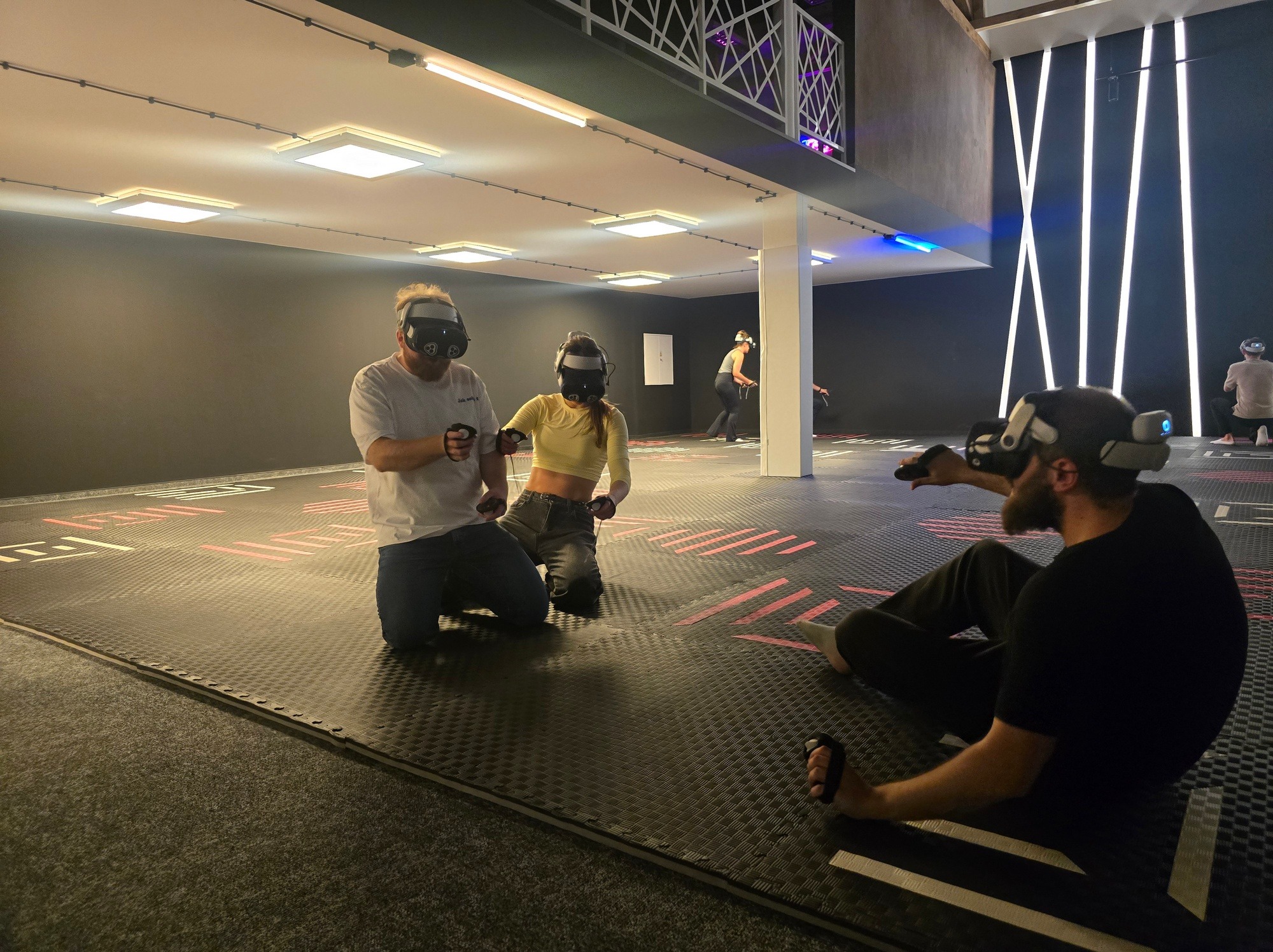 Grupa osób w goglach VR, grających w wirtualną rzeczywistość w nowoczesnym, oświetlonym pomieszczeniu z neonowymi akcentami i wzorzystą podłogą.