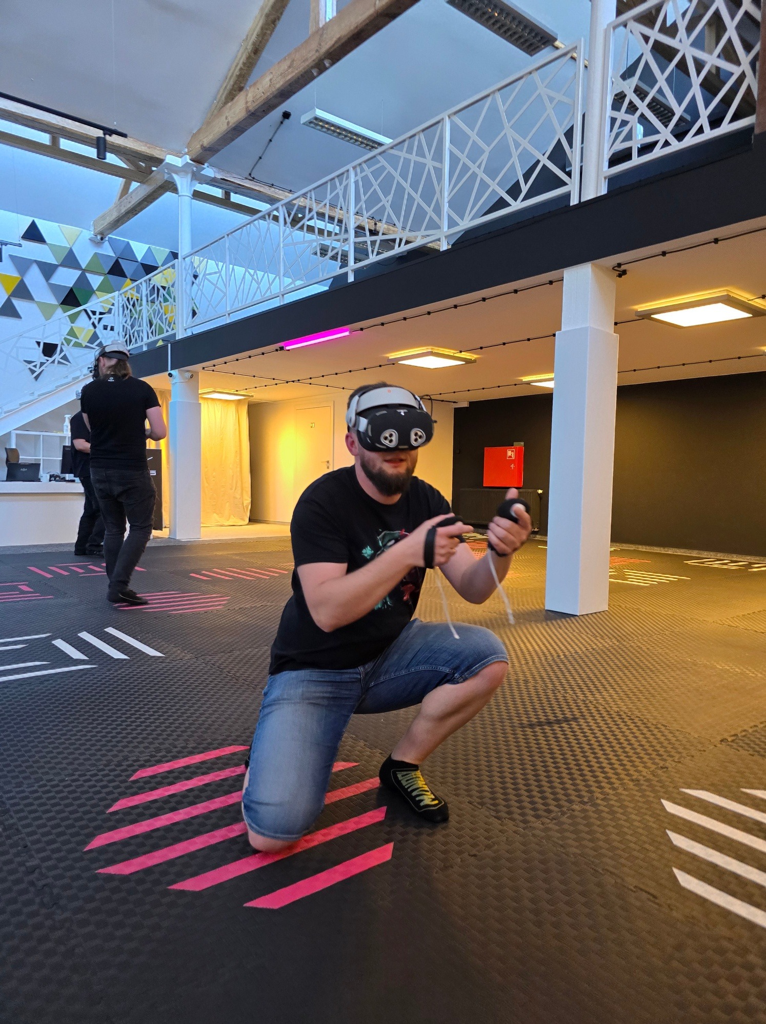 Mężczyzna w okularach VR kuca na arenie z neonowymi pasami, trzymając kontrolery. W tle inni gracze i industrialna przestrzeń eventowa z antresolą.