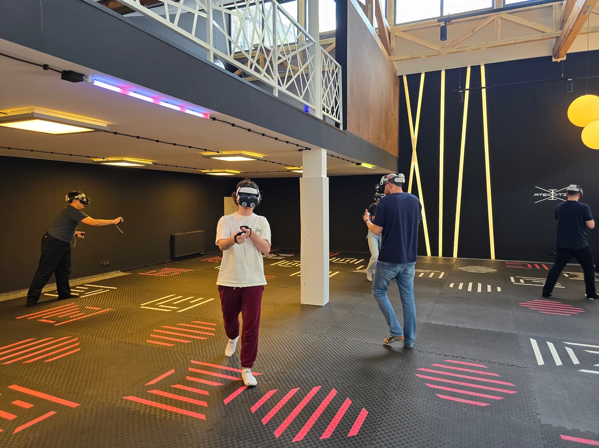 Grupa osób z goglami VR w przestronnym pomieszczeniu z podłogą w geometryczne wzory, czarnymi ścianami z neonowymi paskami i antresolą z białymi balustradami.