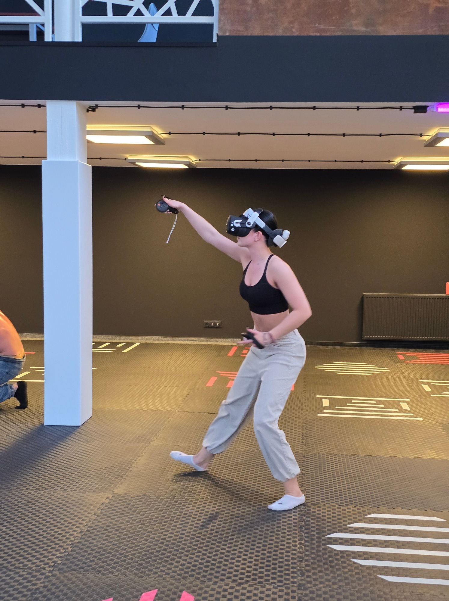 Kobieta w goglach VR, z kontrolerami w dłoniach, w przestrzeni z oznaczeniami na podłodze, sugerującymi wirtualną rzeczywistość i interaktywną rozrywkę.