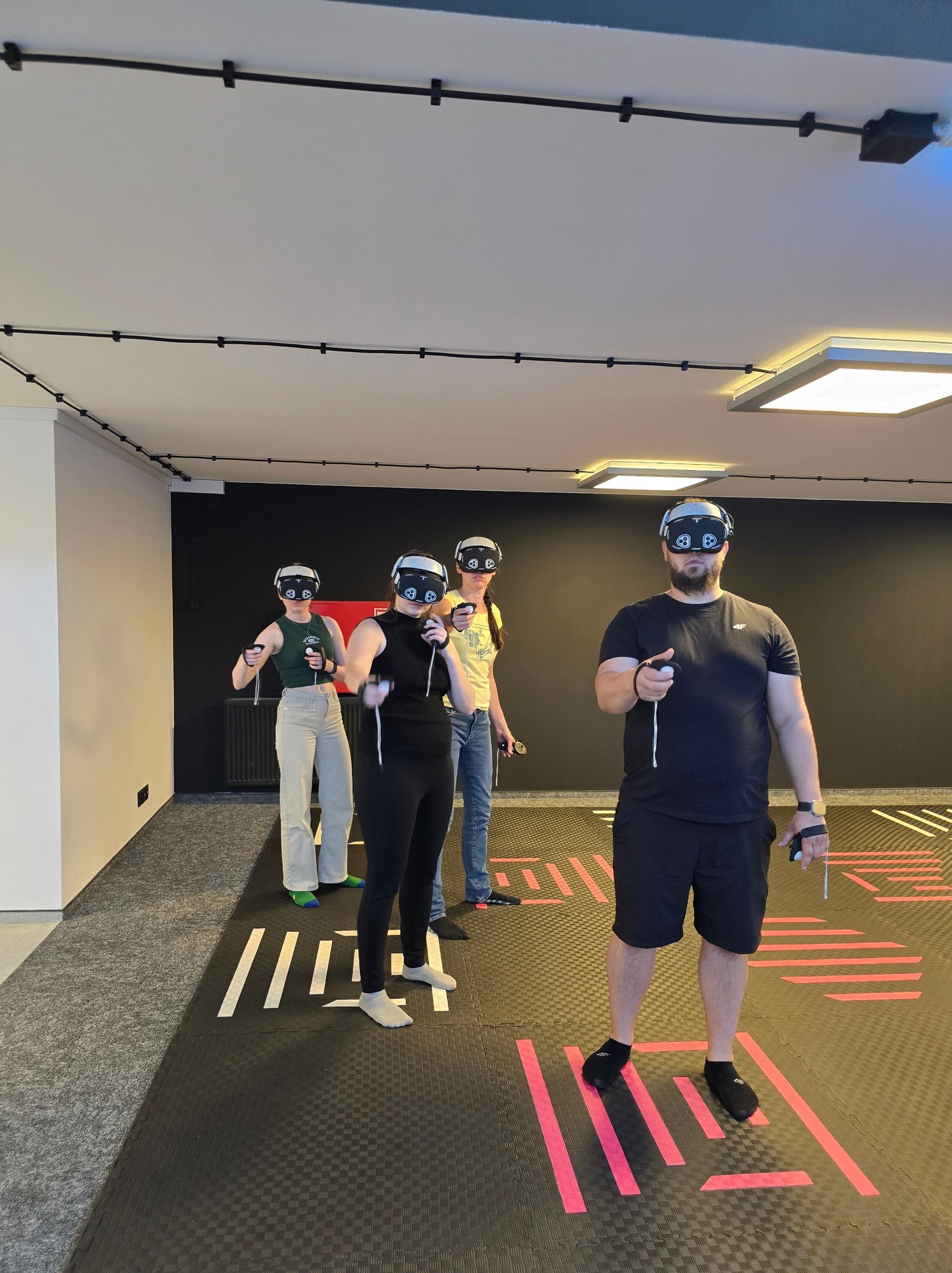 Grupa czterech osób z goglami VR i kontrolerami w dłoniach, na czarnej podłodze z różowymi liniami, w jasnym pomieszczeniu z czarną ścianą. Aktywność w wirtualnej rzeczywistości.