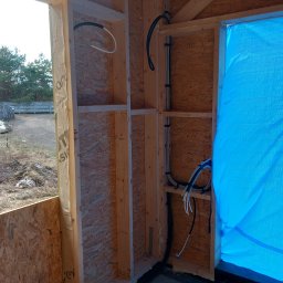 WOJTEX Wojciech Pikuła - Wnętrze szkieletowej konstrukcji domu w trakcie budowy z widocznymi przewodami elektrycznymi przymocowanymi do ściany z płyty OSB. Częściowo widoczne okno i niebieska folia.