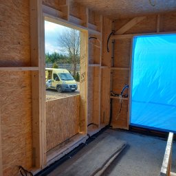 WOJTEX Wojciech Pikuła - Wnętrze szkieletowej konstrukcji domu w budowie z widocznymi przewodami elektrycznymi w ścianach i podłodze. Na zewnątrz żółty bus.