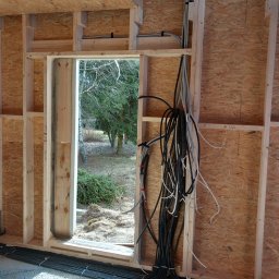 WOJTEX Wojciech Pikuła - Instalacja elektryczna w surowym budynku: kable prowadzone w ścianach z płyty OSB, widoczne przewody pod podłogą i przez otwór okienny. Etap prac przed wykończeniem.