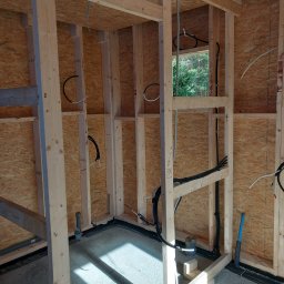 WOJTEX Wojciech Pikuła - Surowy szkielet drewniany z widocznymi przewodami i rurami instalacji wewnątrz ścian. W tle okno z widokiem na zieleń. Widać etap prac instalacyjnych w nowym budynku.