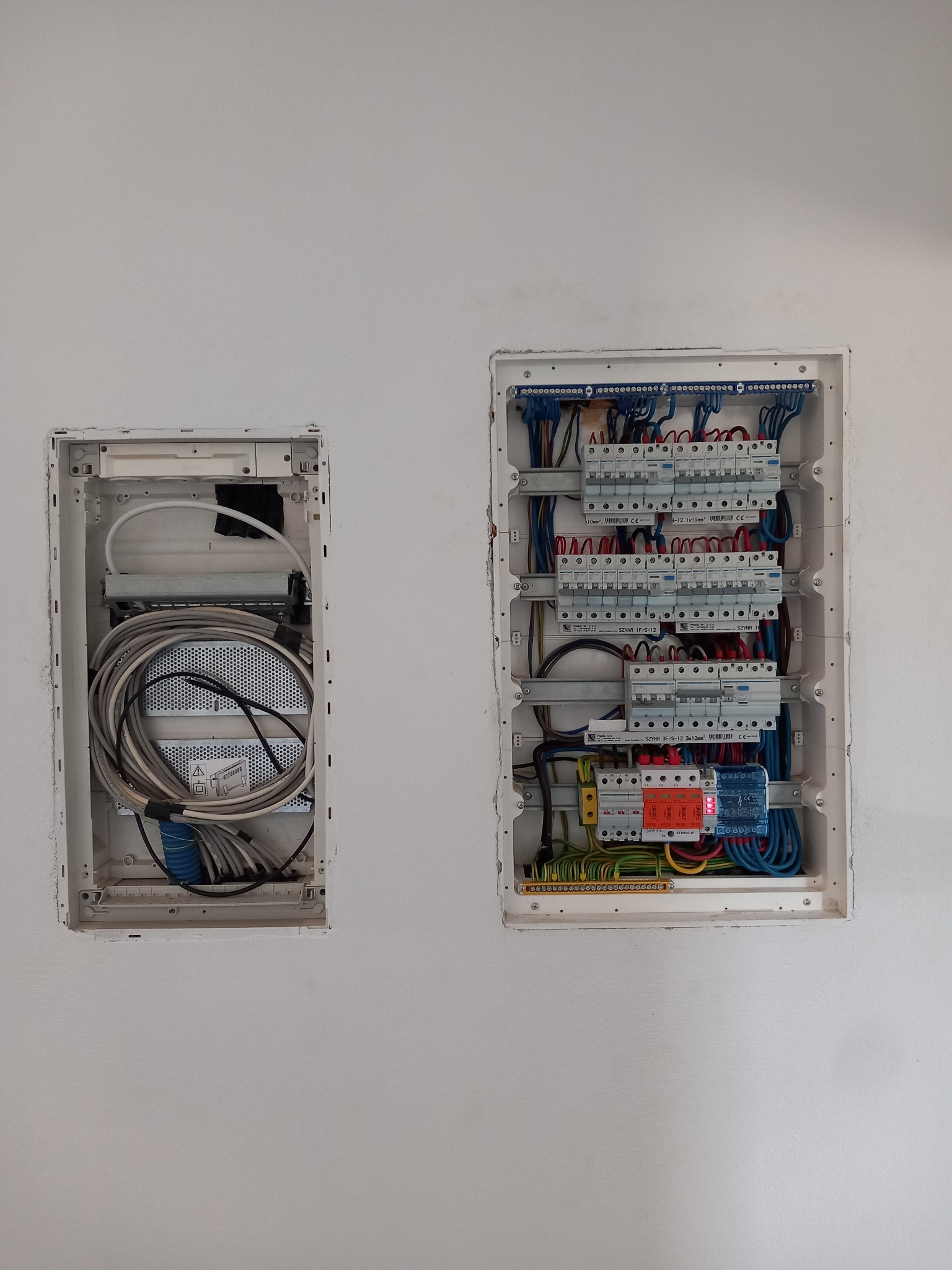 Dwie podtynkowe skrzynki: rozdzielnica z bezpiecznikami i okablowaniem oraz skrzynka z kablami telekomunikacyjnymi. Białe tło ściany.
