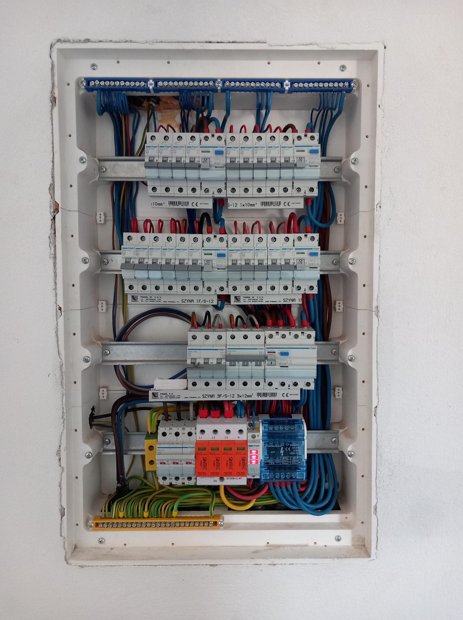 Rozdzielnica elektryczna z zamontowanymi bezpiecznikami, przewodami i listwami zaciskowymi w białej obudowie, osadzona w ścianie. Widoczne oznaczenia i kable w różnych kolorach.